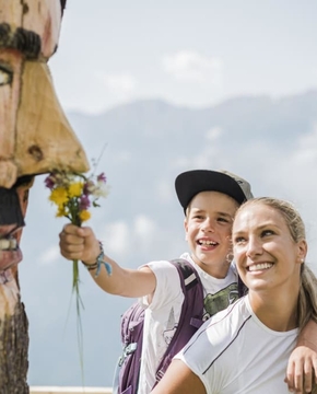 Wanderurlaub mit geschnitzter Holzmaske Ein Kind hält einer lächelnden Frau auf einer Bergwanderung einen kleinen Blumenstrauß hin, neben einer geschnitzten Holzmaske.