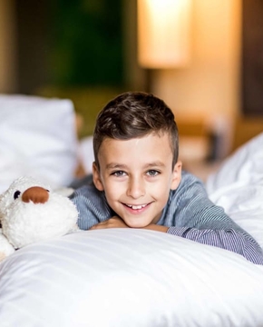 Familienzimmer im Hotel in Naturns Ein Junge liegt lächelnd auf einem Bett und hält ein Kuscheltier im Familienzimmer.